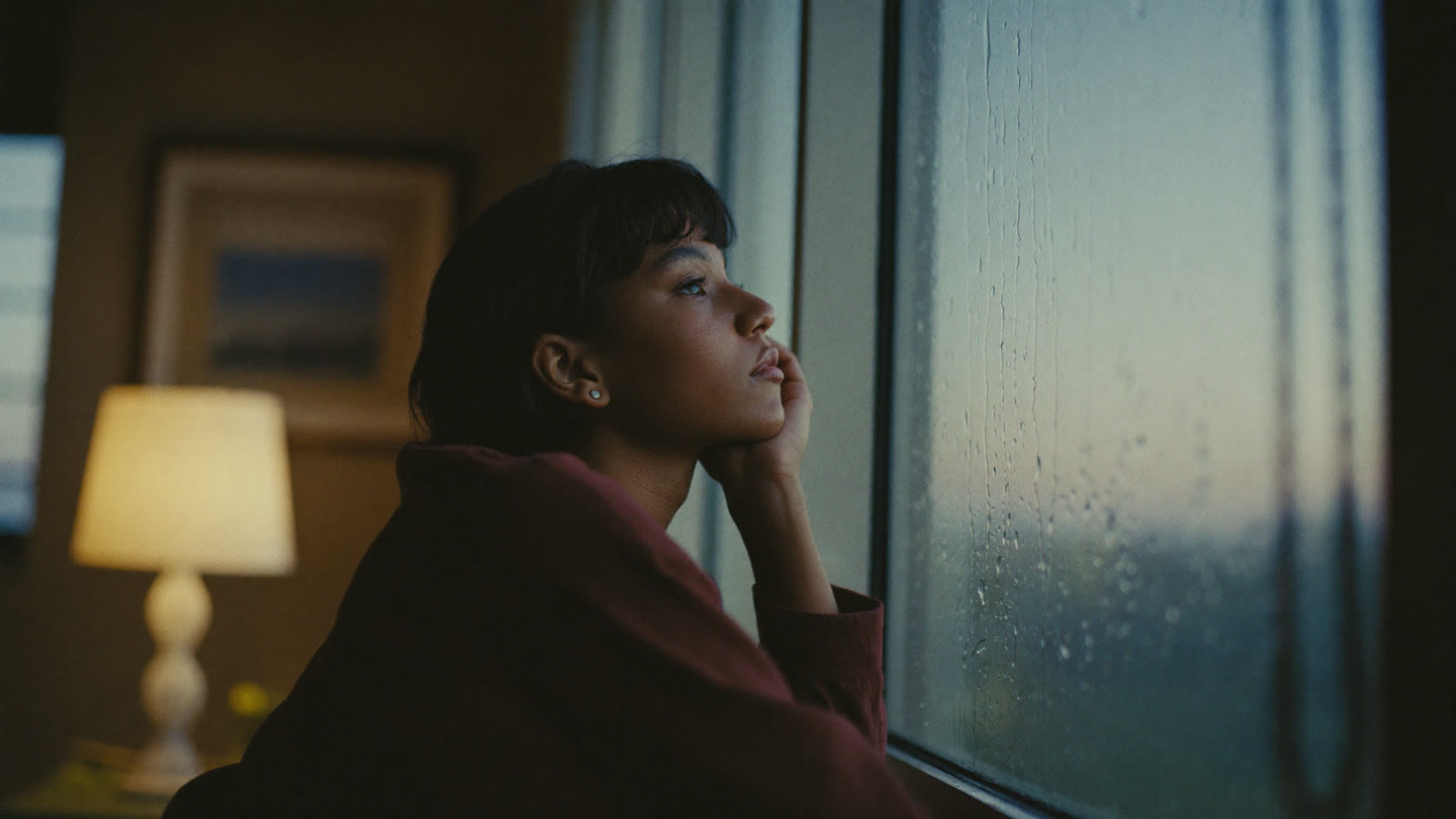 A person gazing softly out a rain-streaked window at dusk, warm lamp glow behind